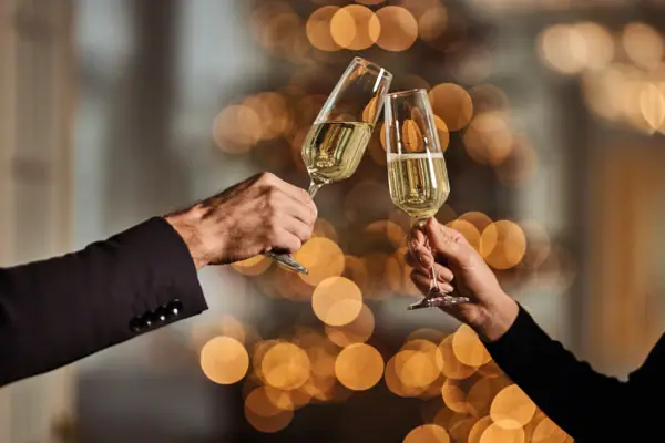 Zwei Hände stoßen mit Sekrgläsern an vor verschwommenen Lichtern im Hintergrund.