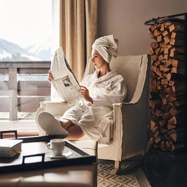 Eine Frau im weißen Bademantel liest eine Zeitung in einem Schaukelstuhl.