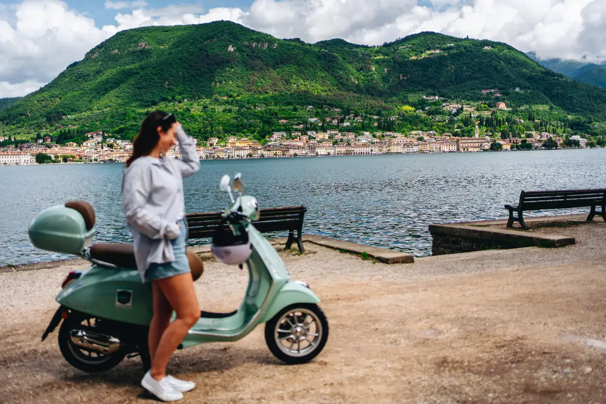 Vespa Gardasee Eine Frau steht neben einem Roller an einem See.