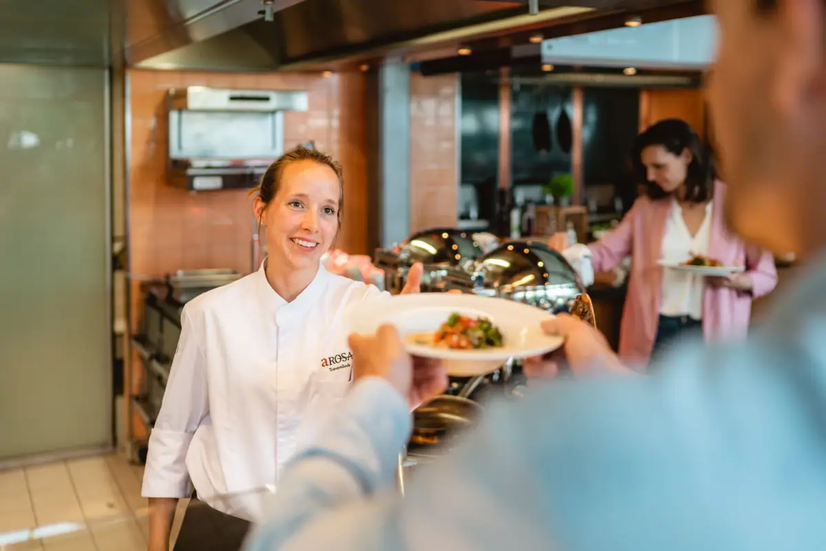 Buffet Travemünde Eine Frau in weißer Kochjacke reicht einem Mann einen Teller mit Essen.