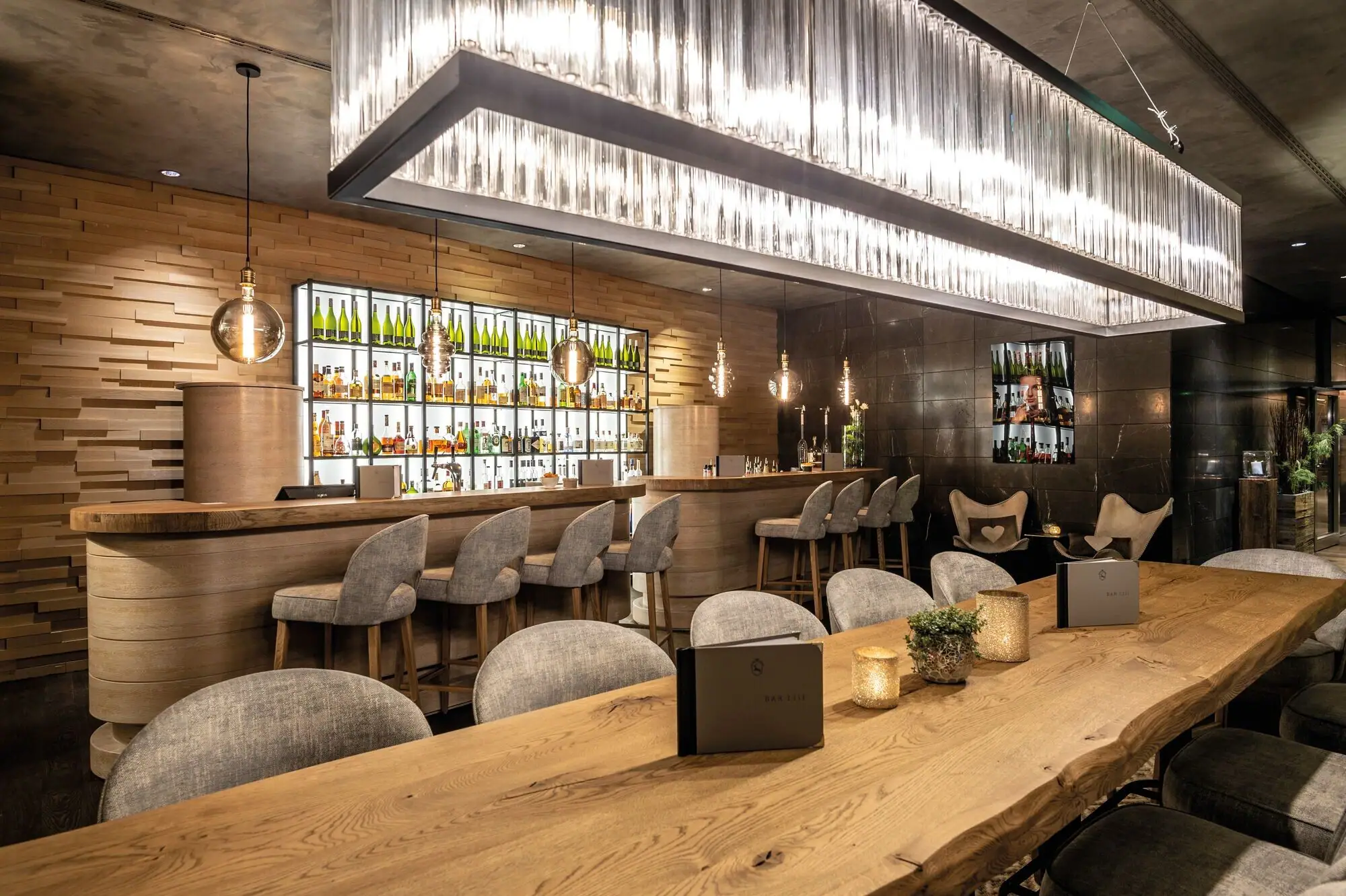 Bar 1.111 im Ifen Hotel Kleinwalsertal Ein langer Holztisch mit Stühlen vor einem Bartresen mit Barhockern vor einer gut gefüllten Bar im Hintergrund..