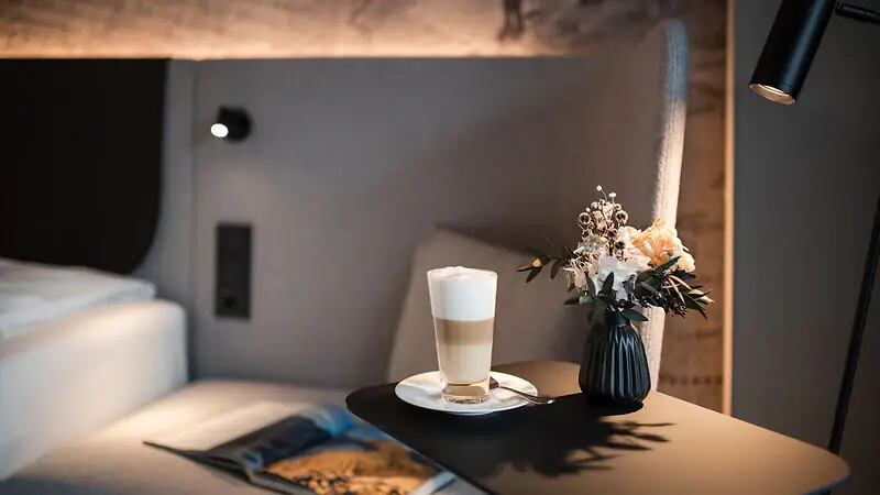 Zimmer & Suiten im Ifen Hotel Kleinwalsertal Ein Beistelltisch mit einem Latte Macchiato, einer aufgeschlagenen Zeitschrift und einer Vase mit Blumen neben einem Bett mit gepolstertem Kopfteil vor einer kunstvoll gestalteten Tapete..