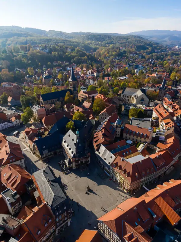 Luftaufnahme von Wernigerode Luftaufnahme einer Stadt mit Häusern und Bäumen, umgeben von Bergen und Wolken am Himmel.