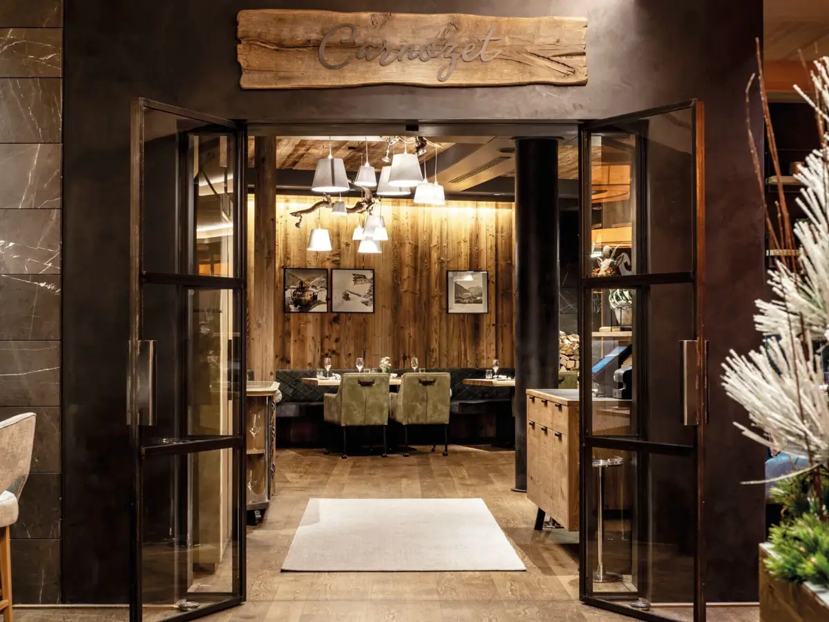 Eingangsbereich Carnozet Ein gemütlicher Restaurantbereich mit vielen Holzdetails in Form von dem Boden sowie Holzwänden. Die Stühle und Bänke sind in natürlichen Grüntönen gehalten, an den Wänden hängen Bilder und weiße Lampenschirme, die von der Decke hängen, spenden Licht.