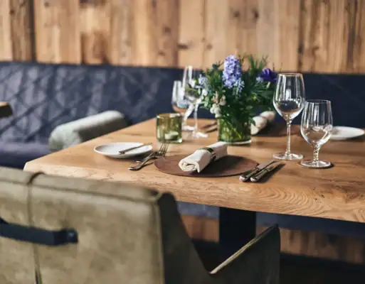 Gastronomie im Ifen Hotel Kleinwalsertal Ein rustikaler Holztisch gedeckt mit Weingläsern und Servietten sowie Blumendekoration und einer gepolsterten Sitzbank vor einer hölzernen Wand im Hindergrund..