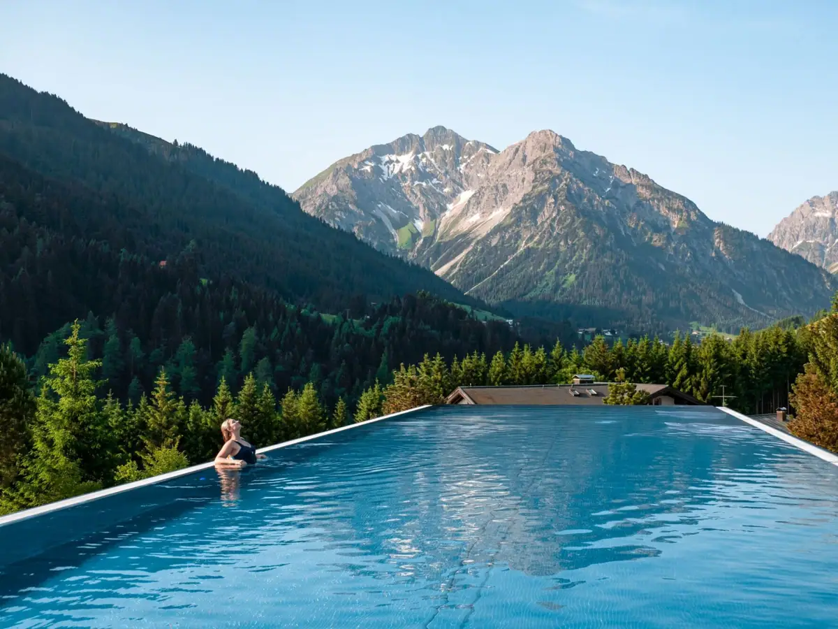 Outdoor Infinity Pool im Ifen Hotel Kleinwalsertal Eine Frau lehnt am Beckenrand in einem Infinity Pool unter freiem Himmel mit Bergen im Hintergrund.
