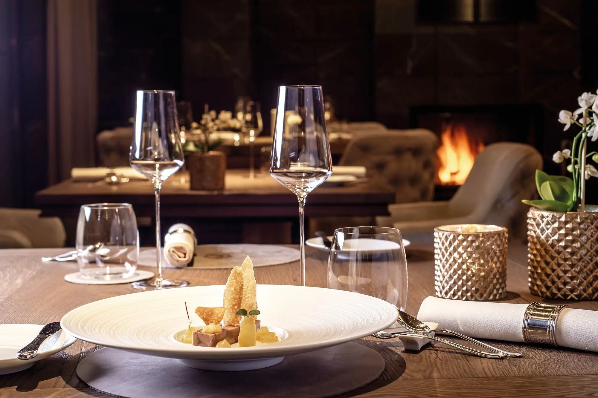 Sternerestaurant Kilian Stuba im Ifen Hotel Kleinwalsertal Ein Tisch mit einem exquisiten Gericht auf einem Teller und Weingläsern in einem Innenraum vor einem Kaminfeuer im Hintergrund..