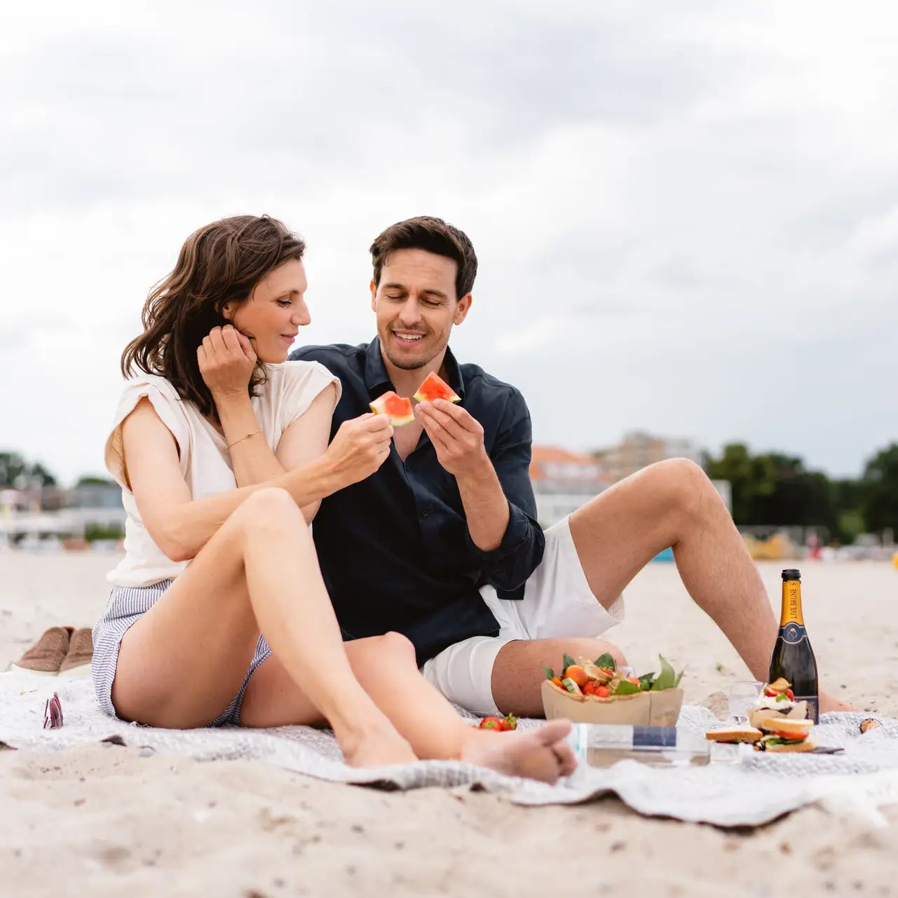 Ein Mann und eine Frau sitzen auf einer Decke am Strand und essen.