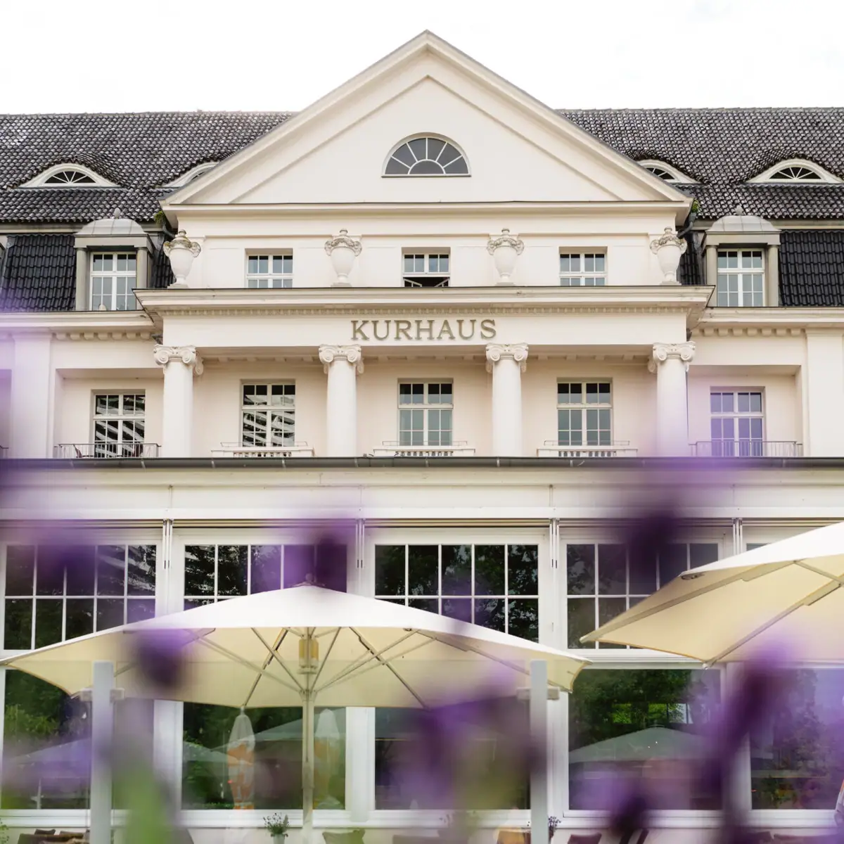 Das A-ROSA Travemünde mit einer großen Fensterfront und einem großen, weißen Sonnenschirm davor.