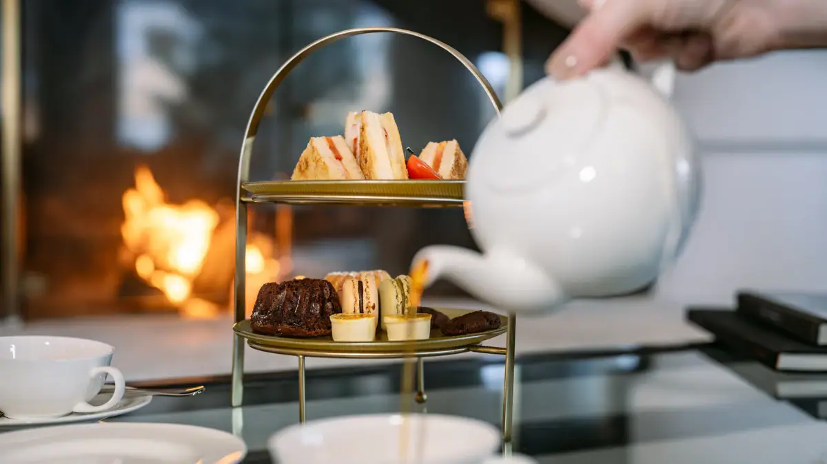 Afternoon Tea Teekanne gießt Tee in eine Tasse im Hintergrund ist eine Etagere mit Essen.