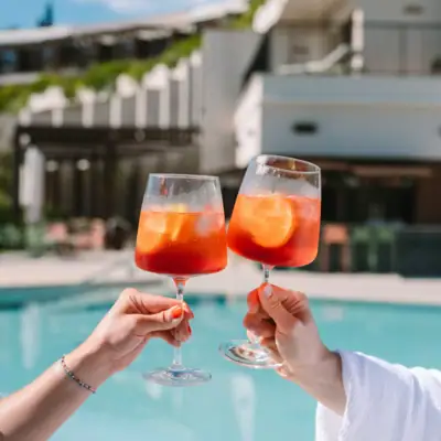 Poolbar Ein Paar Hände hält Weingläser mit orangefarbener Flüssigkeit und Eis vor einem Außenpool im Hintergrund..