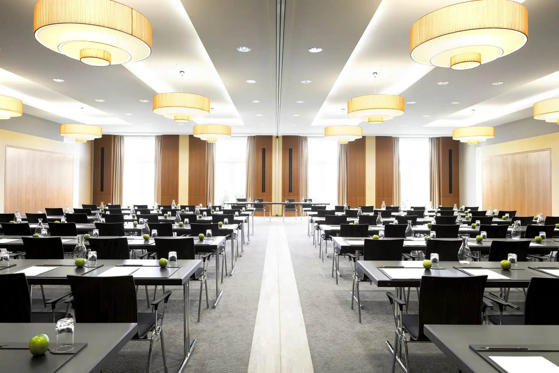 Seminarraum Großer, heller Seminarraum im A-ROSA Hotel auf Sylt mit Reihen von schwarzen Stühlen und Tischen, vorbereitet für eine Konferenz oder Schulung. Auf jedem Tisch liegen ein Notizblock und ein Stift, flankiert von je einem Wasserglas und einem grünen Apfel. Elegante, runde Deckenleuchten hängen über dem Raum und sorgen für eine gleichmäßige, warme Beleuchtung. Große Fenster am Ende des Raums bieten Tageslicht und eine Aussicht nach draußen.