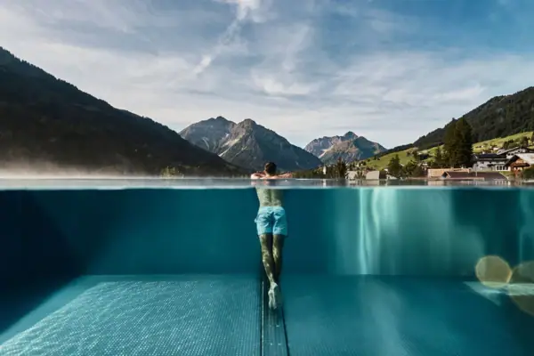 Ein Mann lehnt am Rand eines Outdoor Infinity Pools mit Bergen im Hintergrund.