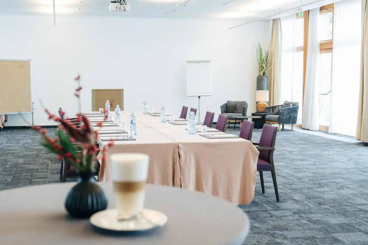 Tagungsraum A-ROSA Ifen Hotel Kleinwalsertal Im Vordergrund ist ein Stehtisch mit einer Blume sowie einem Kaffee zu seheen, während im Hintergrund ein Tagungsraum ist mit einer Blocktafel, um die mehrere Stühle stehen.