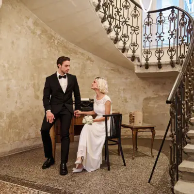 Ein Mann im Anzug und eine Frau im Hochzeitskleid sitzen auf einem Stuhl in einem Raum mit Treppe.