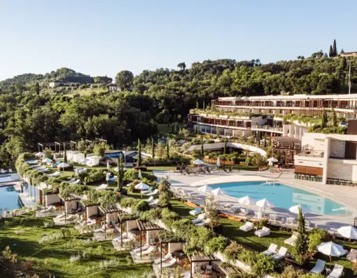 Schwimmbecken mit angrenzendem Gebäude, umgeben von Bäumen und Hügel im Hintergrund.