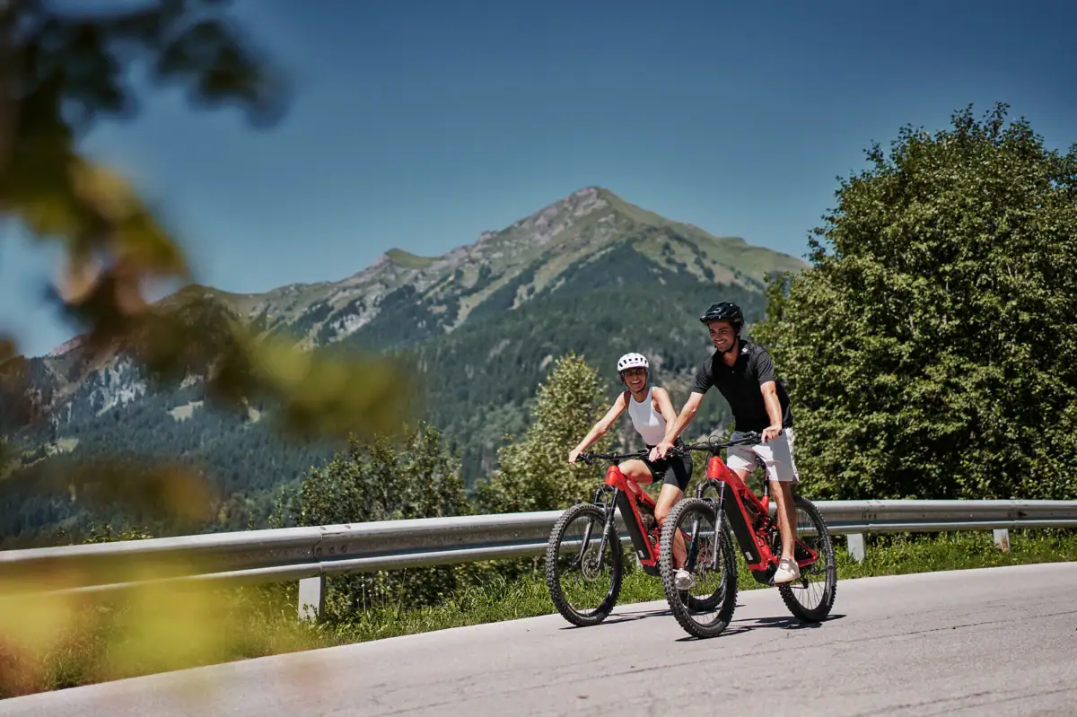 Ein Mann und eine Frau fahren auf Fahrrädern auf einer Straße mit Bergen im Hintergrund.