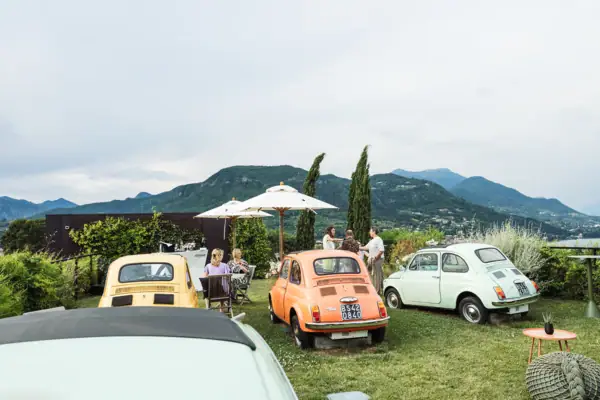 Die Terrasse 500 Eine Gruppe von Fiat 500 Autos auf dem Rasen geparkt, umgeben von Menschen, die sitzen und stehen. Sie halten jeweils Getränke in der Hand. Sonnenschirme sind aufgestellt. Im Hintergrund sieht man die italienischen Berge.