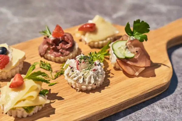 Canapés Kurhaus Binz Fein garnierte Canapés auf einem Holzbrett, belegt mit Käse, Roastbeef, Frischkäse, Schinken und frischen Kräutern, angerichteten als Fingerfood bei Sonnenschein.