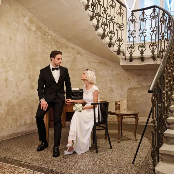 Ein Mann im Anzug und eine Frau im Hochzeitskleid sitzen auf einem Stuhl in einem Raum mit Treppe.
