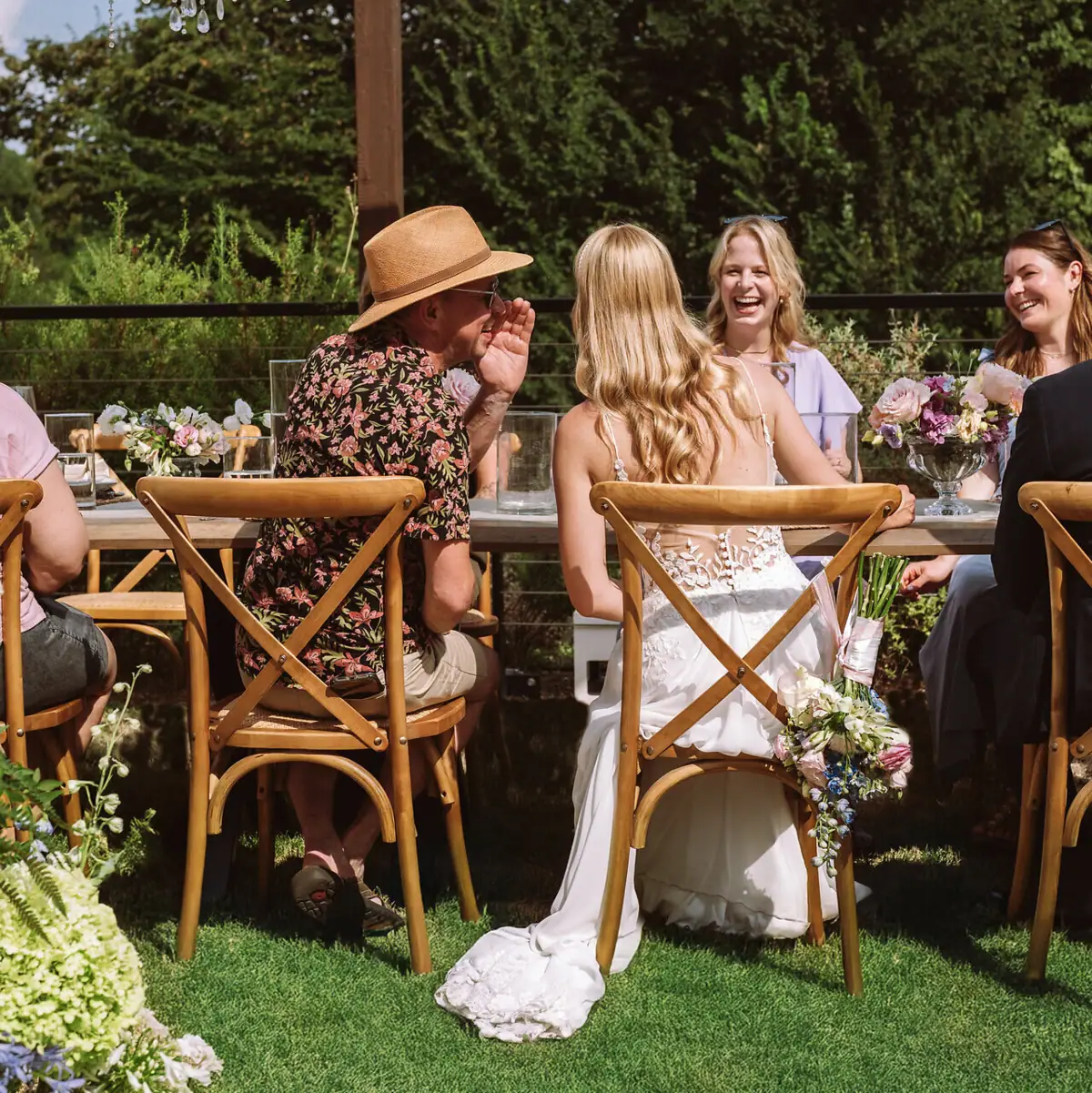 Hochzeit A-ROSA Gardasee Eine Hochzeitsgesellschaft sitzt an einem Tisch im Freien und der Brautvater flüstert der Braut etwas in Ohr.