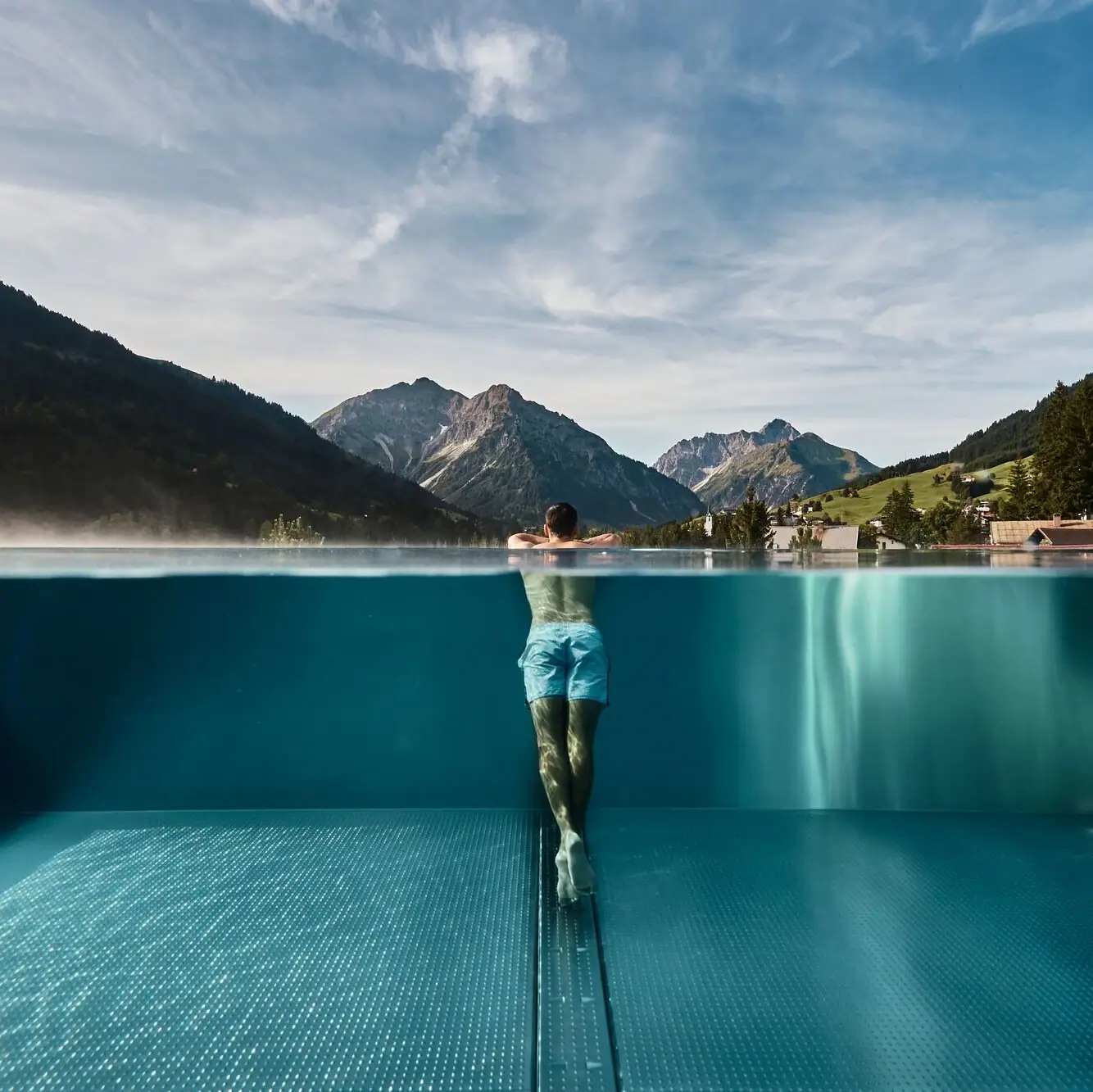 Wellness im Kleinwalsertal Mann lehnt am Rand eines Infinity-Pools mit Blick auf einen Ort inmitten einer grünen Berglandschaft im Hintergrund.