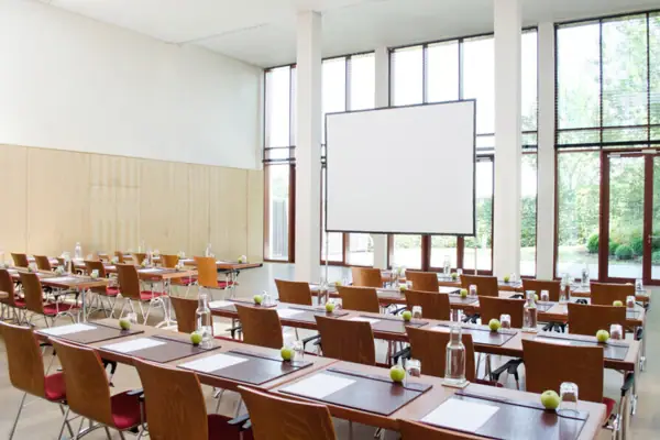 Tagungsloft Ein Tagungsraum mit Tischen und Stühlen vor einer großen Glasfront, wodurch viel Tageslicht in den Raum kommt.