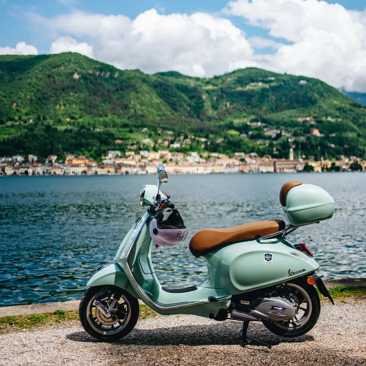 Vespa-Tour am Gardasee Eine mintgrüne Vespa steht am Ufer des Gardasees, im Hintergrund die malerische Uferstadt und die grünen Hügel der Region.