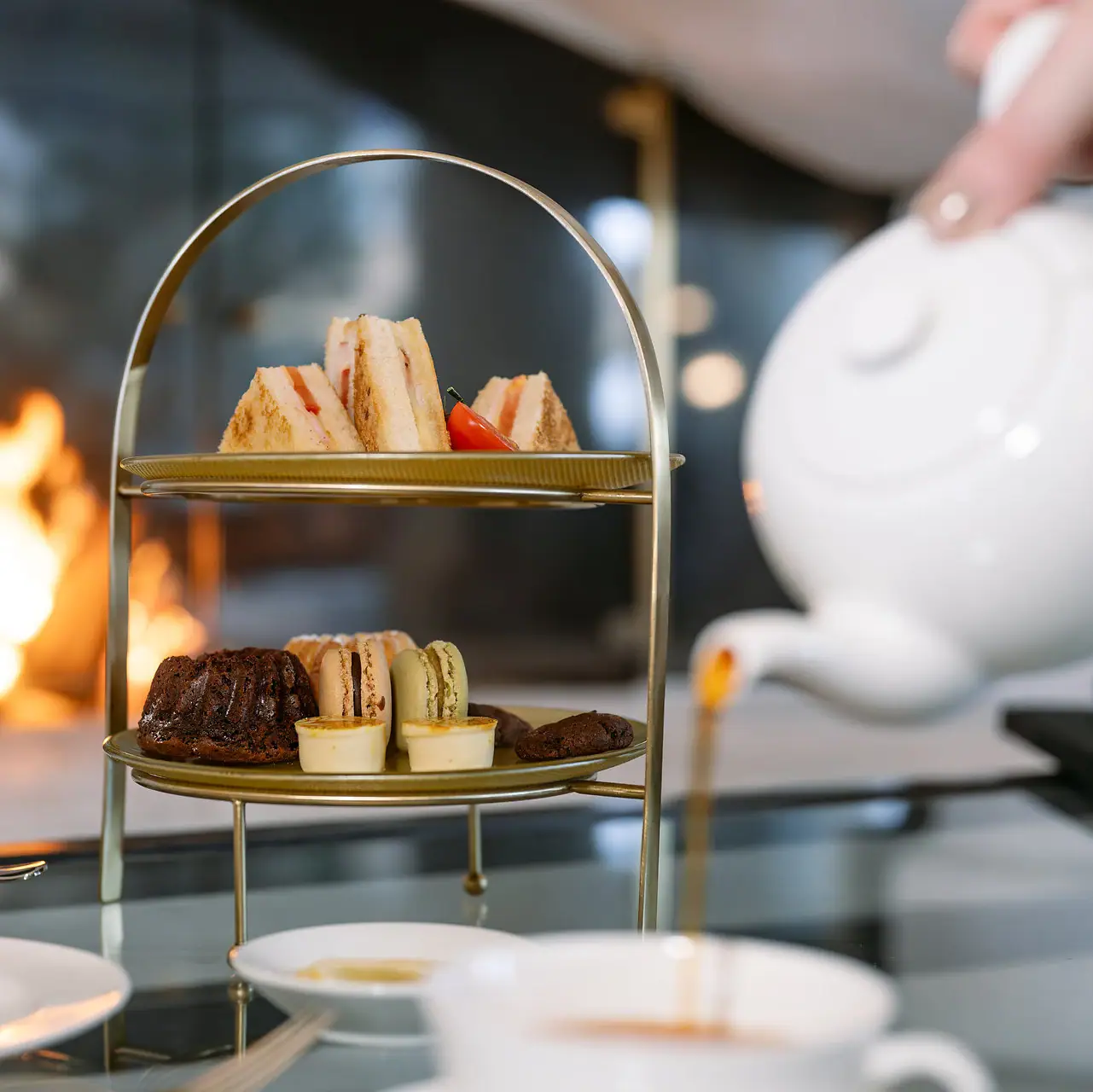 Thurnher's Alpenhof Teekanne gießt Tee in eine Teetasse vor einer Etagere mit Küchlein und einem Kaminfeuer im Hintergrund.