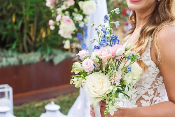 Strahlende Braut mit Hochzeitsstrauß Eine Braut im Spitzenkleid hält einen farbenfrohen Hochzeitsstrauß mit Rosen, Lisianthus und blauen Akzenten.