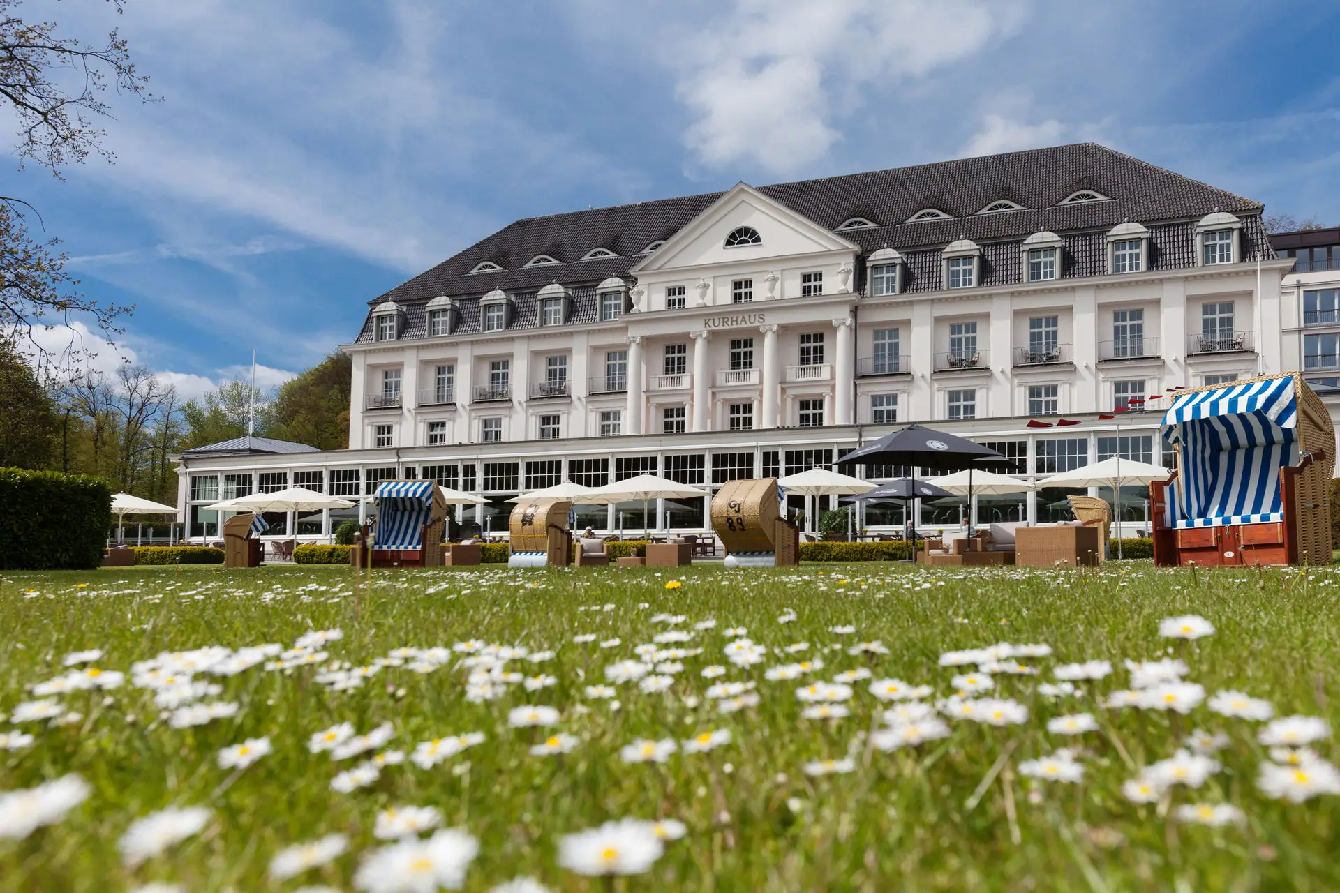A-ROSA Travemünde Rasen mit Stühlen und Sonnenschirmen vor einem großen weißen Gebäude.