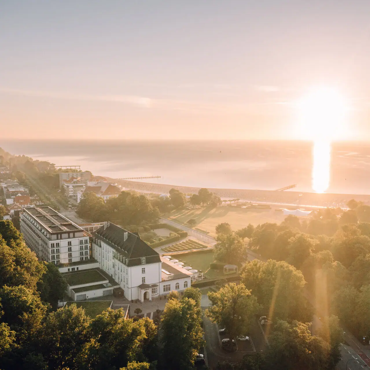 A-ROSA Travemünde Angebote Ein großes Hotelgebäude umgeben von Bäumen vor einer Küste bei Sonnenaufgang.