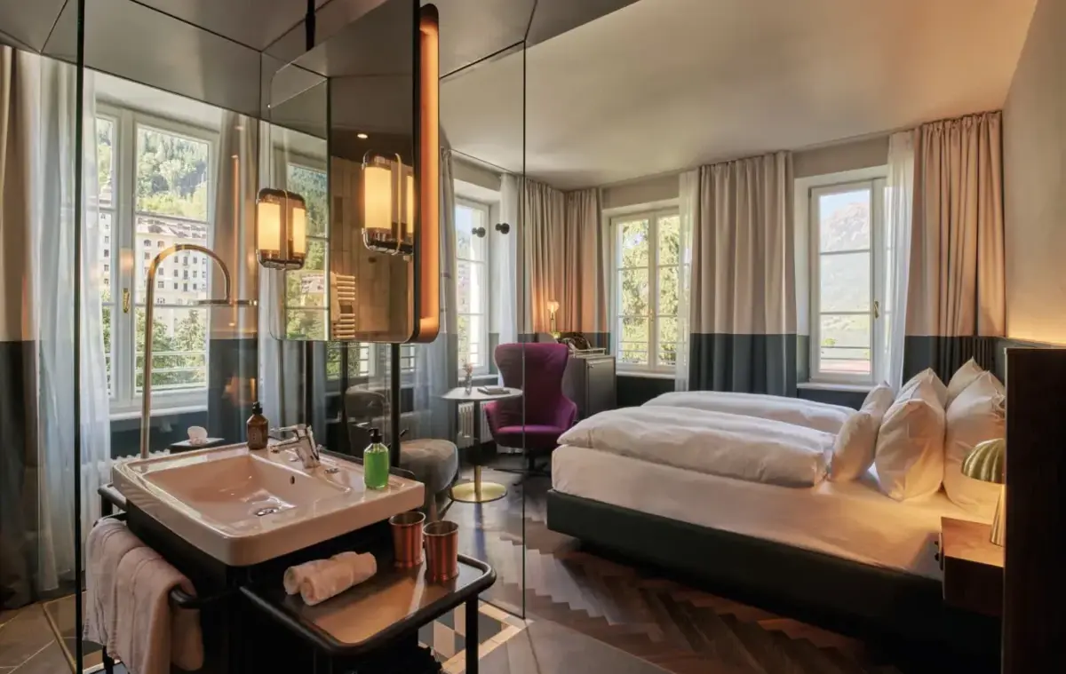 Individuell gestaltete Zimmer, so einzigartig wie unsere Gäste Blick in ein Hotelzimmer mit verglastem Badbereich, Doppelbett und Sitzgelegenheit am Fenster.