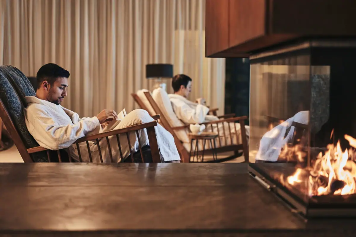 Ein Mann sitzt in einem Sessel und liest ein Buch vor einem Kamin.