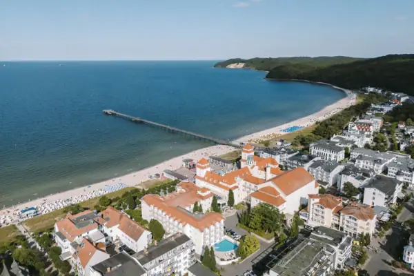 Außenansicht Kurhaus Binz Die Küste von Binz aus der Luft fotografiert mit einem Strand, Gebäuden und einem Pier bei blauem Himmel.