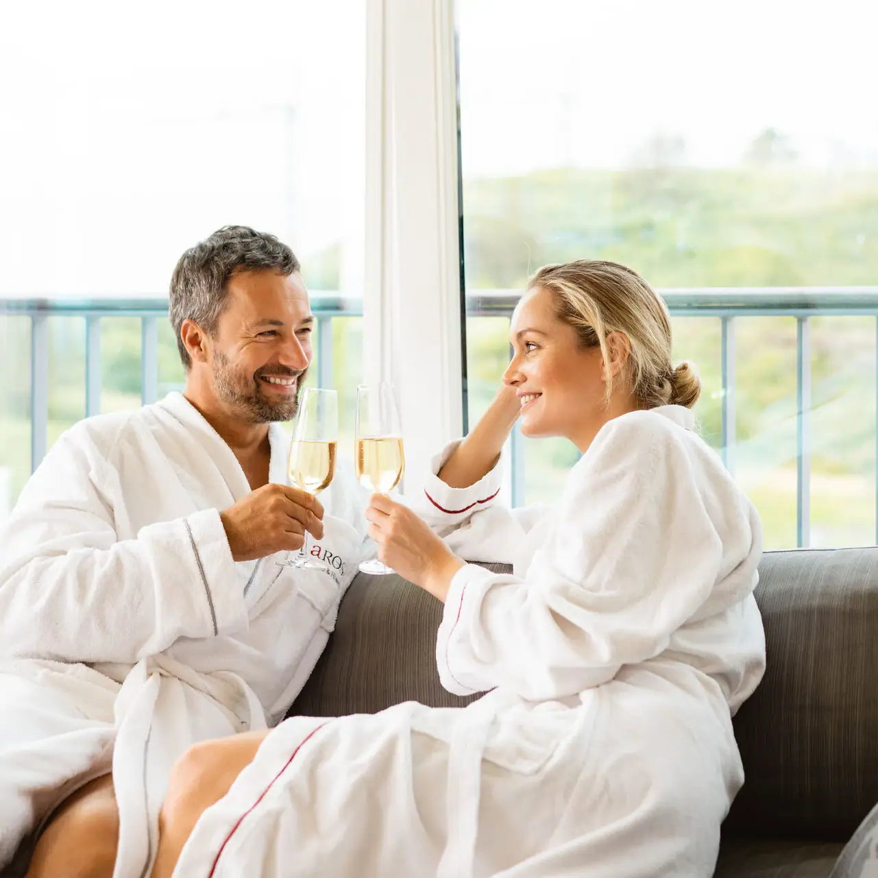 Ein Mann und eine Frau in Bademänteln sitzen auf einem Sofa und halten Champagnergläser.