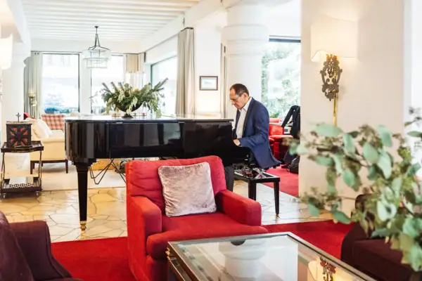 A-ROSA Hotel in Zürs Ein Mann im Anzug spielt Klavier in einem Raum mit roten Stühlen und einem Tisch.