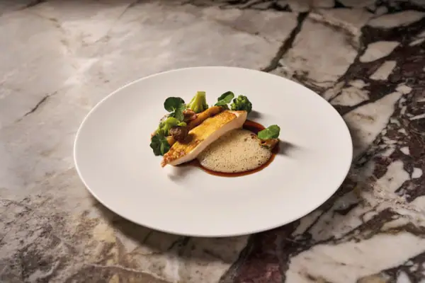 Essen als Kunstwerk inszeniert. Ein weißer Teller mit einem Gericht darauf steht auf einem marmorierten Untergrund.