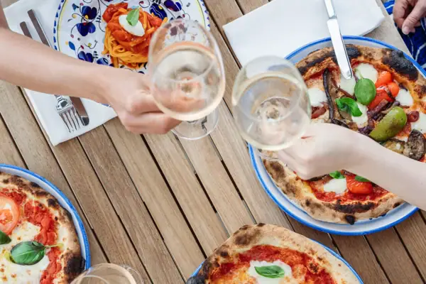 Geselligkeit bei Pizza und Drinks Hände stoßen mit Weingläsern an, daneben frisch gebackene Pizza auf einem Holztisch.