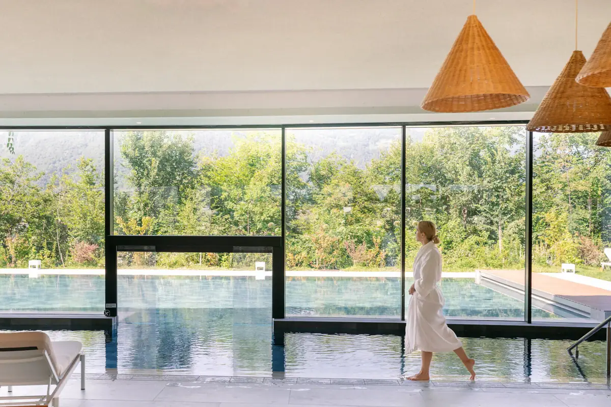 Innen- und Außenpool im SPA-ROSA Gardasee Frau im Bademantel spaziert am Innenpool des A-ROSA Gardasee entlang, mit Blick auf den Außenpool und die umliegende Natur.