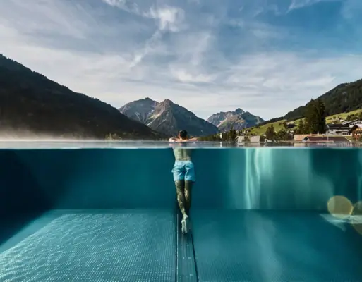 Ein Mann lehnt am Rand eines Outdoor Infinity Pools mit Bergen im Hintergrund.