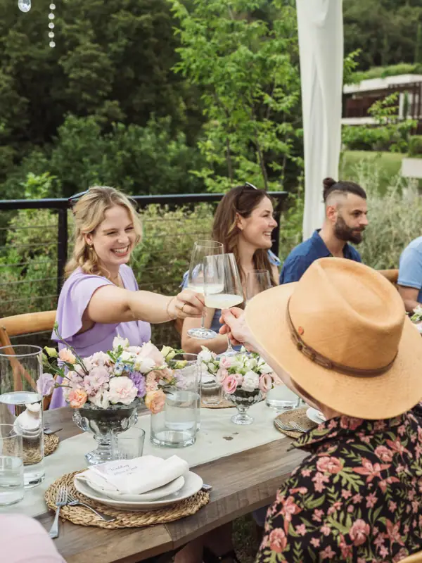 Fröhlicher Abend auf der Hochzeit Hochzeitsgäste sitzen an einem gedeckten Tisch im Freien und stoßen lachend mit Gläsern an.