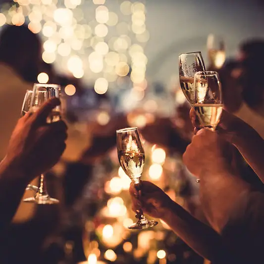 Eventhighlights Mehrere Hände halten Sektgläser in die Höhe vor verschwommenen Lichtern im Hintergrund.