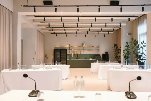 Creavista im A-ROSA Gardasee Ein Tagungsraum mit Tischen und Stühlen. Der vordere Tisch ist mit Mikrofonen sowie Gläsern und Wasserflaschen eingedeckt. Die hinteren Tische sind auf mit Gläsern und Wasserflaschen eingedeckt. Ganz hinten im Raum sieht man einen Kühlschrank und eine Küchenzeile für eine Kaffeepause.