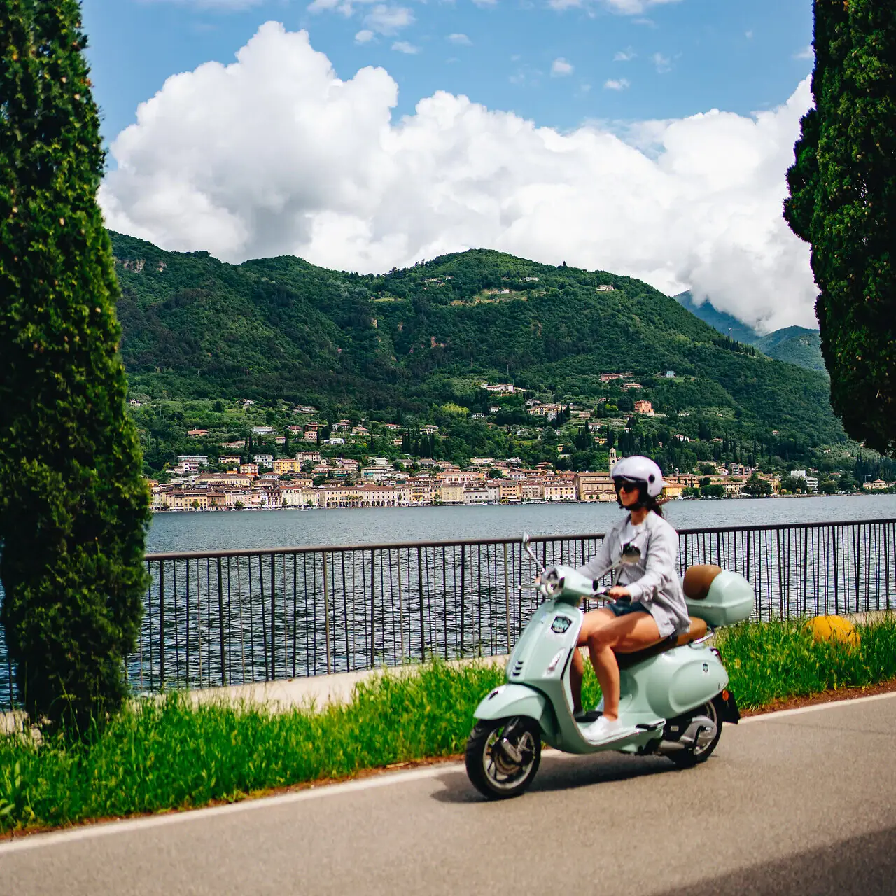 Eine Frau fährt mit einem Roller auf einer Straße an einem Gewässer entlang.