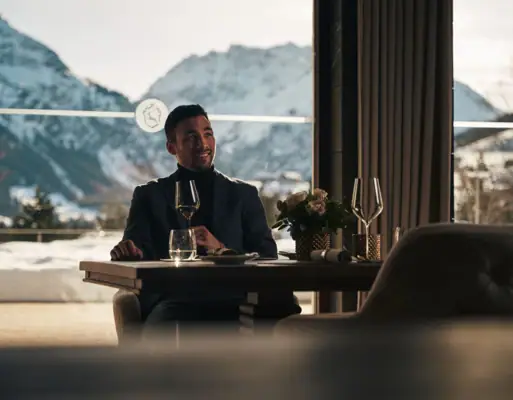 Ein Mann sitzt an einem Tisch mit Weingläsern und Bergen im Hintergrund.