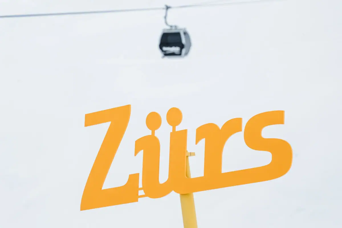 Gelbes Schild vor Seilbahn im Hintergrund, umgeben von schneebedeckten Bergen.