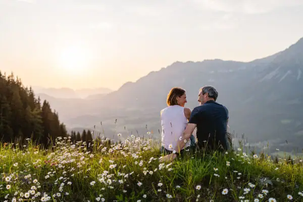 Ein Mann und eine Frau sitzen auf einem Hügel mit Blumen und Bergen im Hintergrund.