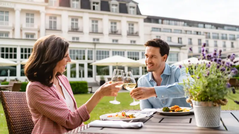 Terrasse Travemünde Ein Mann und eine Frau stoßen mit Weingläsern an einem Tisch im Freien an.