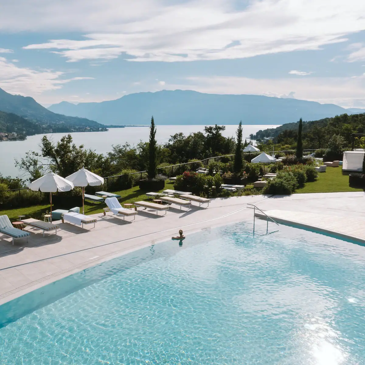 Pool mit Panoramablick auf den Gardasee Außenpool im A-ROSA Gardasee mit Blick auf den See und die umliegenden Berge, umgeben von Liegen und Sonnenschirmen.